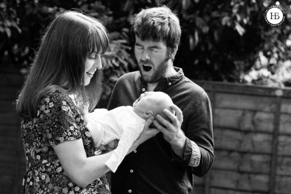 Two parents with their newborn baby photographed outdoors in their garden during a home newborn session. Father and baby yawn in unison while mother laughs.