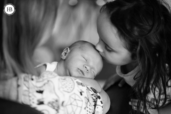 Newborn baby sleeping in soft natural light during an at-home photography session, older sister softly kisses their young siblings head.