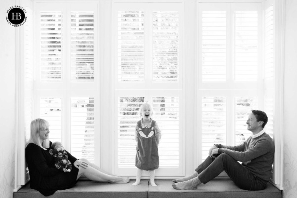 A family sit in a bay window, mum has a newborn baby on her lap and the toddler stands laughing in the centre while dad looks on