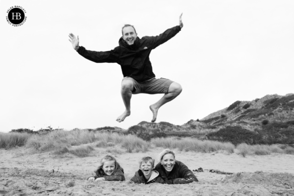 dad jumps over his family who are in a hole dug in the sand on the beach. A fun family photo