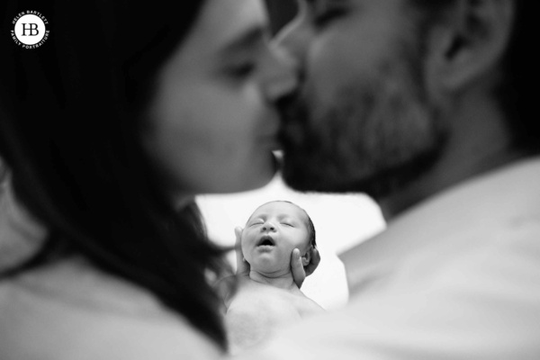 Parents share a kiss while their newborn rests between them — a creative natural family portrait from a London at-home newborn photography session
