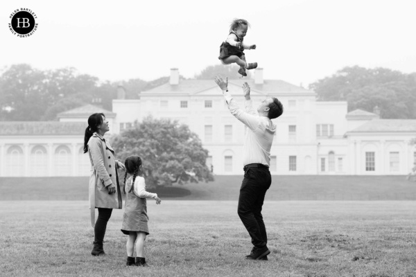 family pictures in front of Kenwood House on Hampstead Heath. Dad throws baby in the air while mum and older daughter look on.