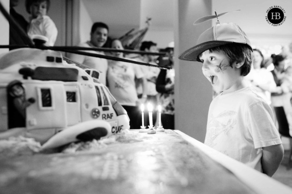 A little boy with a joyful expression and a cap with a propeller on top looks with joy at his fourth birthday cake which is an RAF helicopter. The candles shine brightly in the foreground
