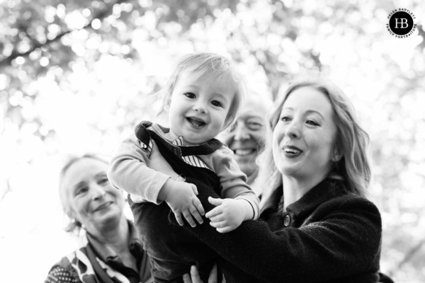 A mother holds her one year old baby to the camera with proud grandparents visible in the background