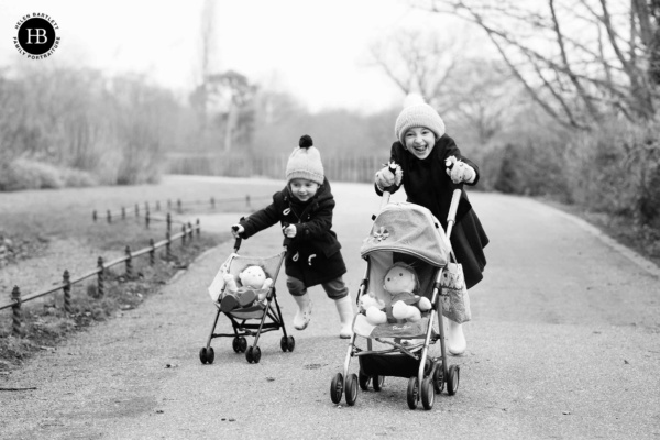 Two children run towards the camera pushing prams with toy dolls in and laughing. They are wearing warm coats and bobble hats in this joyful winter family photography session.
