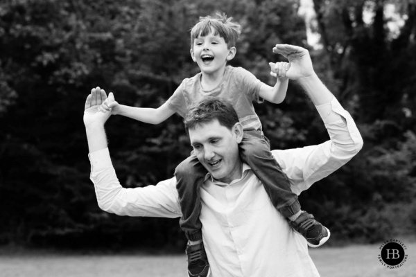 Little boy laughs happily while sitting on his father's shoulders and running through the woods. They hold hands in this joyful father and son photograph.