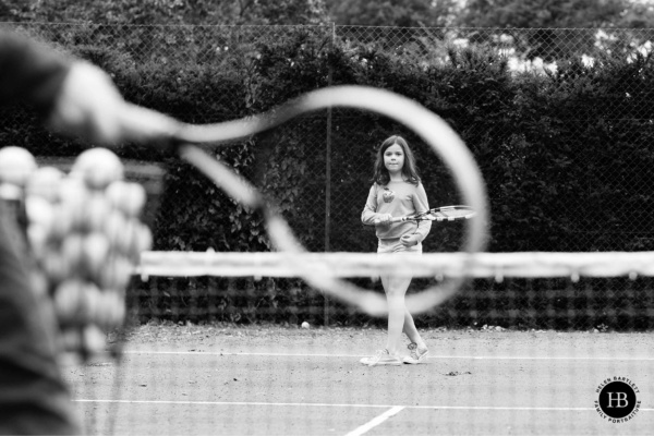 Little girl plays tennis, she is visible through the racket of her older brother in a creative family photo shoot.