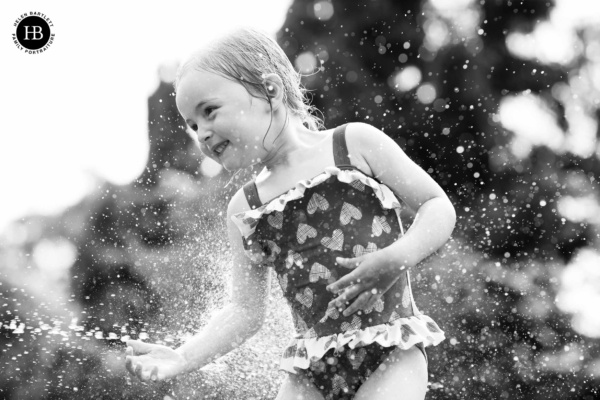 little girl laughing in the spray of a garden hose during a summer family photo shoot with photographer and Canon Ambassador Helen Bartlett