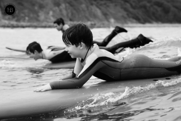 Three boys are surfing in a line. Image taken from in the water by London family photographer Helen Bartlett on a joyful summer family photography session.