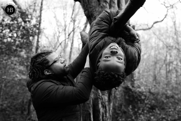 father helps son to hang upside down from a tree during a family photo session. The boy looks at the camera smiling