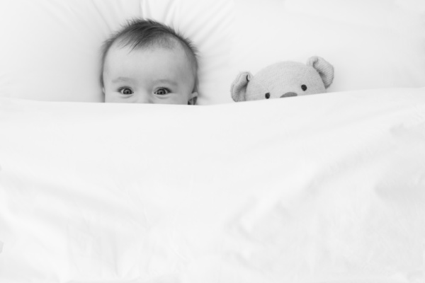 A baby and their bear peep over bedding.