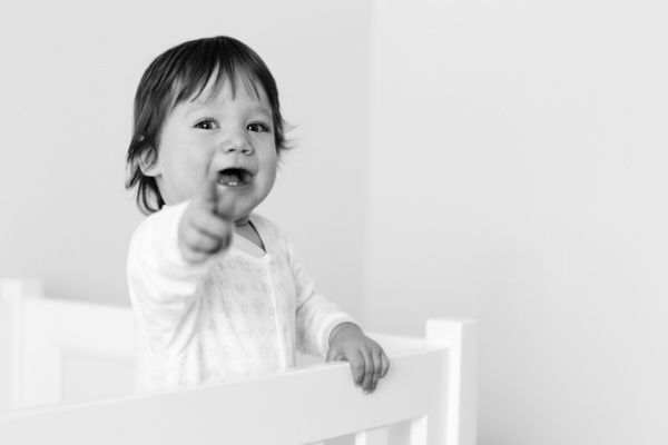 A baby in a white cot points at the camera.