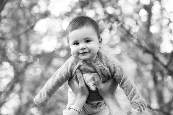 A parent holds a baby in the air with tree branches in the background.