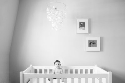 A baby sits in a white cot in a bright nursery.