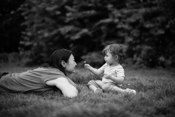 A baby offers their mother a flower while they're sitting on the grass.