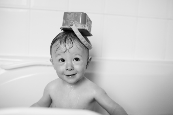 A baby plays in the bath, with a toy on his head.