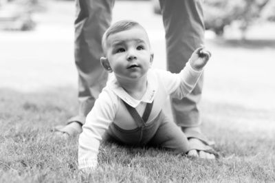 Black and white portrait of a baby crawling near his father's legs.