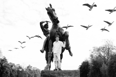 A boy stands in front of a London statue while a flock of geese flies overhead.