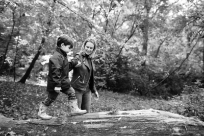 A mother walks her son along a tree branch in woodland.