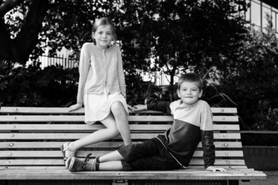 A brother and sister sit on a garden bench for a family portrait.