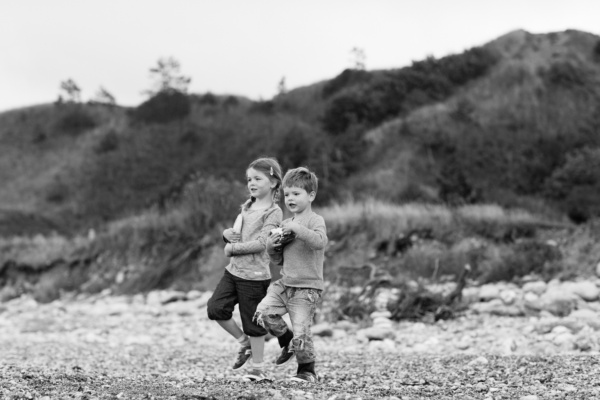 A brother and sister run on a pebbled beach.