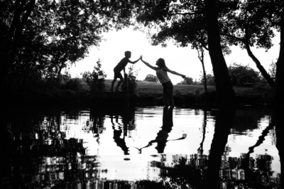 Two children high-five each other near a pond surrounded by trees.