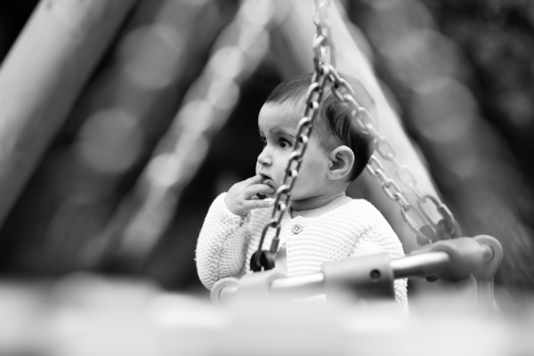 A baby puts their finger in their mouth while at the swings.