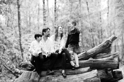 A family of four sits on a pile of logs in woodland.