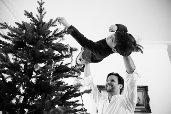 A father lifts his child up to decorate their Christmas tree.