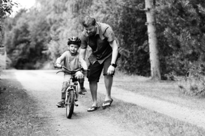 A father helps his son ride a bike along a park.