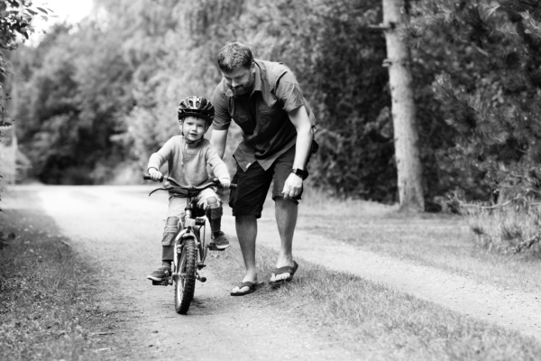 A father helps his son ride a bike along a park.