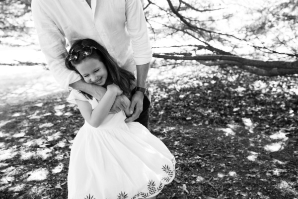 A father hugs his daughter in the shade of a tree.