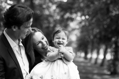 A father and mother laugh with their daughter near a row of trees.