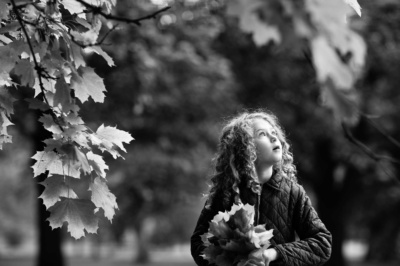 A girl holding a handful of leaves, walks through trees and looks at the sky.