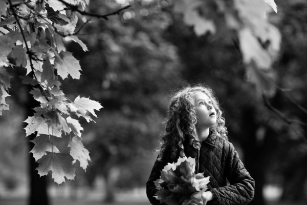 A girl holding a handful of leaves, walks through trees and looks at the sky.