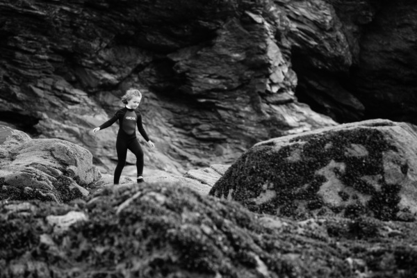 A girl in a wetsuit walks across rocks on a beach.