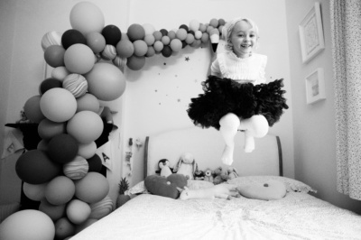 A girl jumps on her bed surrounded by balloons.