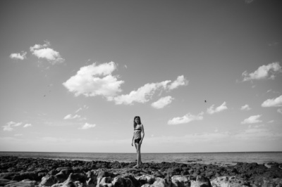 Girl walks along rocky beach with a cloudy sky behind her.