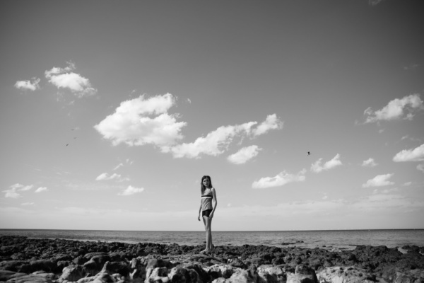 Girl walks along rocky beach with a cloudy sky behind her.