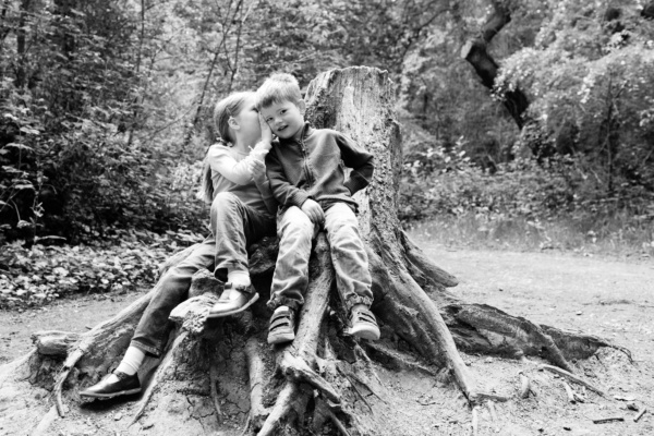 A girl whispers in her brother's ear, while they sit on a log in a clearing.