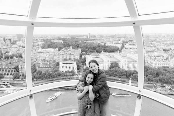 Siblings hug inside one of the London Eye capsules with a panoramic view of London and the Thames in the background.