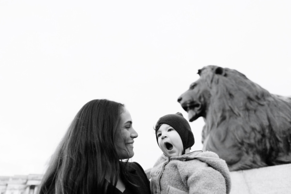 A mother cradles her baby in London's Trafalgar Square, with a lion sculpture in the background.