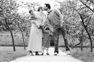 A mother and father kiss in an orchard, while holding hands with their children.