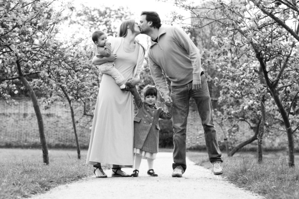 A mother and father kiss in an orchard, while holding hands with their children.