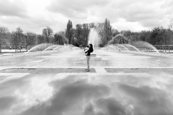 A mother holds her baby near fountains in a park.