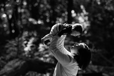 A mother holds up their baby against a background of tree branches.