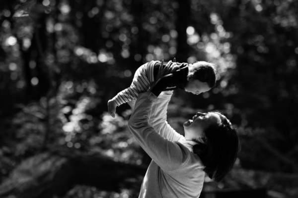 A mother holds up their baby against a background of tree branches.