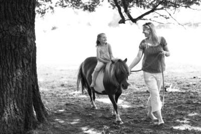 A mother leads her daughter on a pony in the shade of a tree.