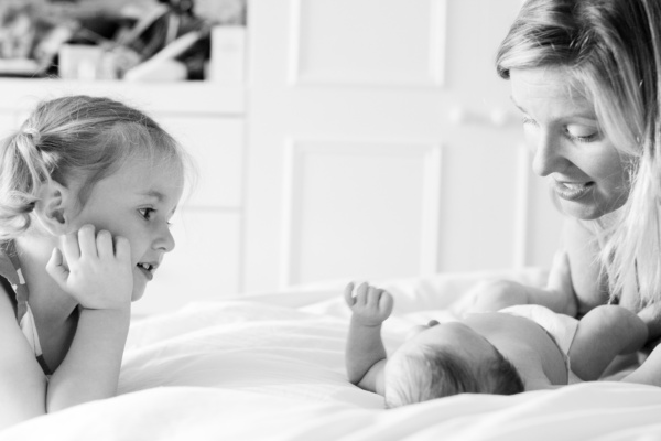 Mother plays with baby on the bed while their sister watches.