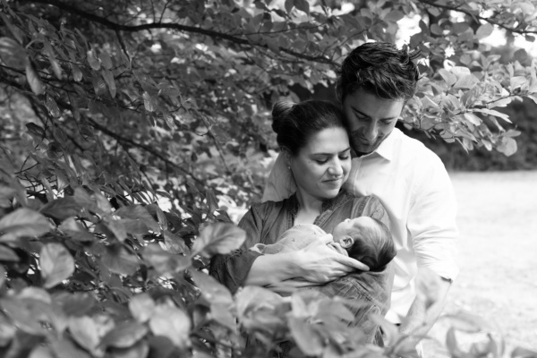 Parents hold their newborn in the shade of a tree.